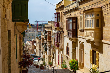 Fototapeta na wymiar Narrow charming street in Cospicua (Bormla), Malta, with stairs and traditional enclosed wooden balconies. Authentic Maltese urban scene.