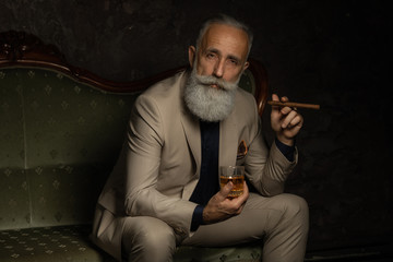 In chair sitting senior business man with cigar and whisky. Gray hair and beard wearing elegant...