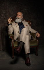 Fototapeta na wymiar In chair sitting senior business man with cigar and whisky. Gray hair and beard wearing elegant suit. Against brown wall.