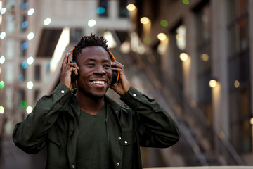 smiling african-american man in stylish jacket in wireless headphones listening music on the street...