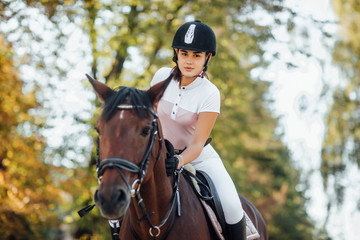 Fototapeta premium Portrait of young jockey girl riding a brown horse in autumn forest.