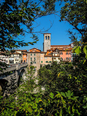 Fototapeta premium Ponte del Diavolo and Bell tower of Cividale del Friuli, Italy. Shot through branches on south shore on sunny day