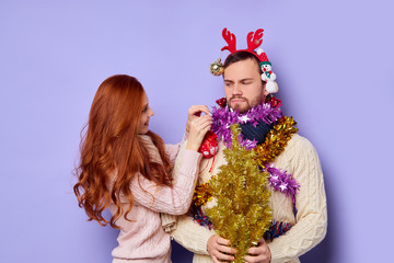 Dissatisfied, displeased man looking at chemung cheerful girl decorating him with different Christmas baubles, isolated family shut, posing over blue background, indoor shot