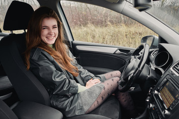 Fototapeta premium Close up portrait of young happy attractive red hair woman driver dressed black leather jacket grey skirt sitting in driver's seat cinematic vintage film filter colors
