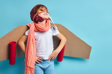 Cute kid wearing plane made from cardboard and pilot mask stand isolated over blue background, looking up, portrait © alfa27