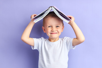 Portrait of smiling kid holding book above head, stand isolated over purple background studio....