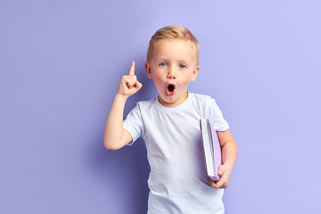 Little boy holding notebook and showing index finger up, portrait isolated purple background