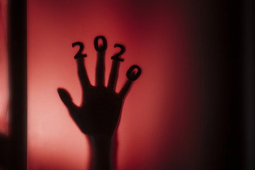 silhouette of a female hand with date on fingers pressed to glass of door with a red strange backlight, destruction of the stereotype happy new year, entering 2020 horror genre