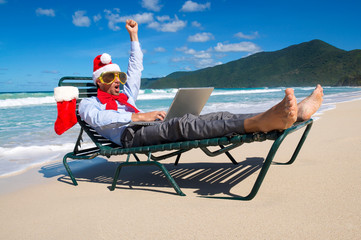 Happy businessman in Santa hat, sunglasses and big red Christmas bow celebrating with a fist pump and his laptop on a tropical beach