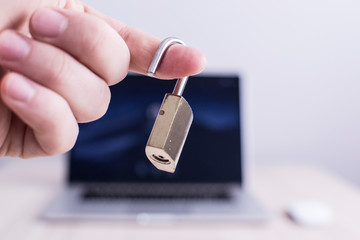 A male hand is holding an unlocked padlock in front of a blurry computer. The padlock is unlocked so it implies also the computer screen is unlocked. Information and data security is important