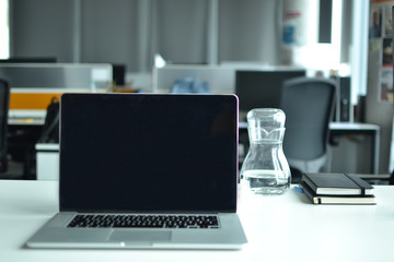 A laptop and monitor on the office desk. Modern furnitures are seem on the back, a carafe and notebooks are also seen on the table