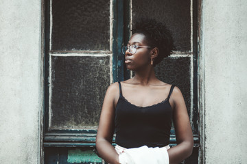 Portrait of a charming black girl in eyeglasses standing in front of a flaked bluish doorway and looking aside; young African female in spectacles, tank top, and with curly Afro hair near the door