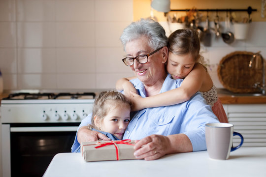 Children congratulate grandmother with gift box. Kids are hugging senior woman in cozy home kitchen. Family enjoying kindness, tenderness. Lifestyle moment. Happy holidays Thanksgiving, Birthday