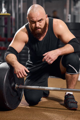 Fototapeta na wymiar Side shot of hairless athlete with thick brunette beard, putting hand on heavy barbell, looking at camera with serious face, posing in gym hall, indoor shot