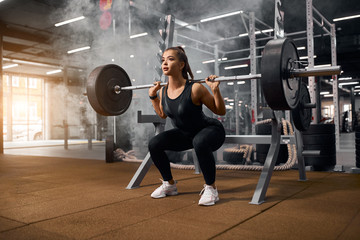 Female weightlifter is getting ready to stand with heavy barbell, squatting with weight, looking away with pleasant expression, enjoying training in gym, portrait, side shot - obrazy, fototapety, plakaty