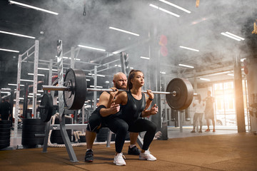 Fototapeta na wymiar Rude brutal trainer with bald head and thick brunette beard standing behind young female powerlifter, backing her up, controlling process of squatting using heavy barbell in gym, white smoke in air
