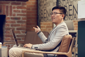 Side view portrait of young Asian businessman using laptop while talking to colleague during meeting in modern office, copy space