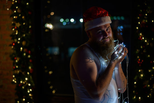 A comic image of santa claus in underwear on christmas eve. A man in a suit of Santa Claus holds a garland with lights in the New Year. A parody of a female photo shoot.