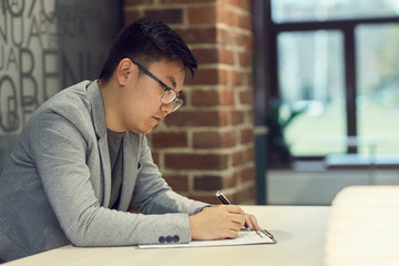 Side view portrait of young Asian businessman writing on clipboard while sitting at desk and...