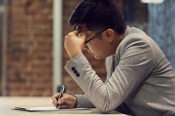 Side view portrait of young Asian man writing on clipboard while working at desk in modern office,...