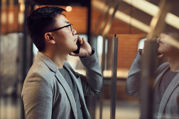 Side view portrait of young Asian businessman speaking by smartphone while standing by glass wall...