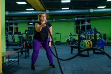 Fat woman doing strength training using battle ropes in the gym. The obese girl is engaged in circular exercises for weight loss.