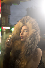 Beautiful caucasian girl  in winter clothes on the street, attractive teenager portrait with a fur jacket , she's watching a timeline of transport on  an electronic screen illuminated by a violet