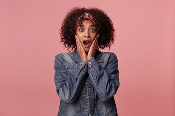 Surprised pretty dark skinned brunette lady with short curly hair holding palms on her face and looking amazedly at camera, rounding eyes with wide mouth opened over pink background