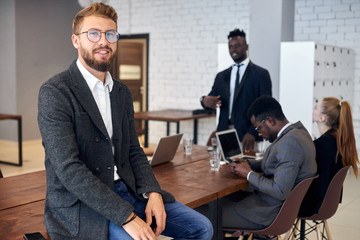Attractive man confidently looking at camera while his business partners have discussion in office