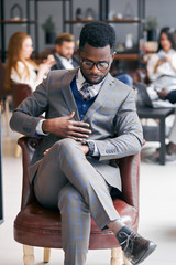 African businessman in grey tuxedo looks at his wristwatch sitting on leather chair while his business colleagues have rest using mobile phones