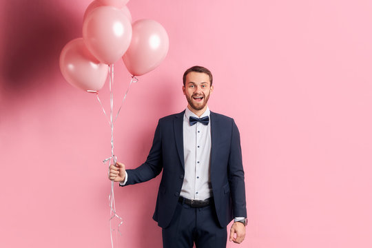 Good-looking man with beard wearing suir stand holding pink air balloons and happily smile, isolated pink background. Celebrating concept