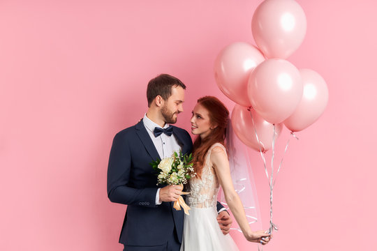 Charming lady with red hair and bearded man prepare to get married with each other, posing isolated over pink background with air balloons.