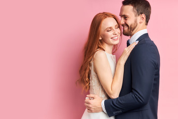 Handsome man in tuxedo and woman in white wedding dress lovely hug each other and smile, after wedding ceremony posing isolated over pink background.