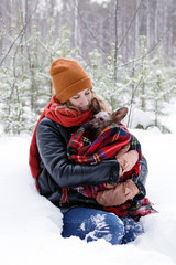 Fototapeta na wymiar Beautiful smiling woman is holding in hands her little dog wrapped in red checkered plaid on a background of winter coniferous forest.