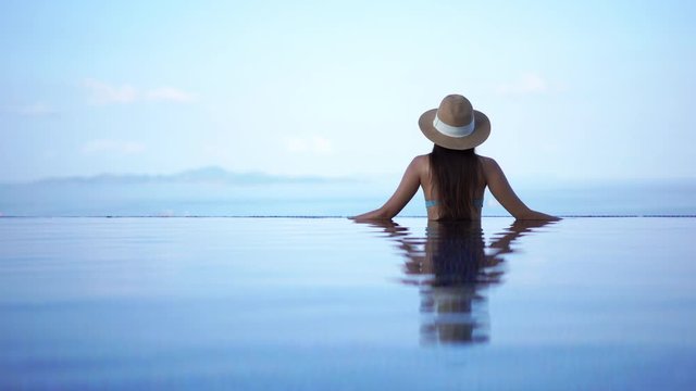 A woman with her back to the camera scans a mountainous island in the tropical ocean waters from the edge of a resort infinity pool.