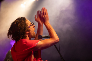 A male musician is viewed from the side as he sings, with open mouth in microphone and clapping...