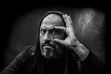 Black and white portrait of a bald bearded man in a hood on a dirty gray background looking at you. Close search concept.