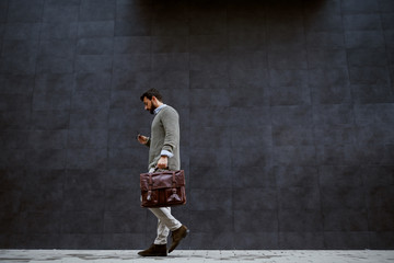 Full length of stylish caucasian designer in gray sweater holding leather bag while passing by gray wall.