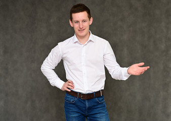 Concept of a pleasant young man manager talking to the camera. Photo Portrait of a secretary guy on a gray background in blue jeans and a white shirt in various poses.