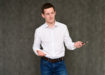 Concept of a pleasant young man manager talking to the camera. Photo Portrait of a secretary guy on a gray background in blue jeans and a white shirt in various poses.