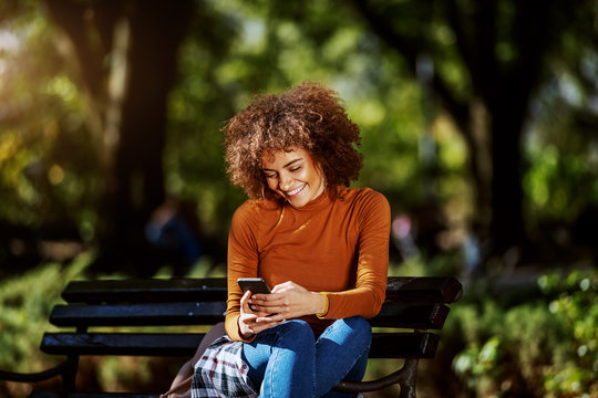 Lovely young smiling mixed race woman in turtleneck and with curly hair sitting on bench in park and using smart phone for reading or sending message.