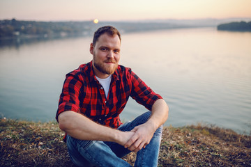 Young handsome bearded caucasian blond man in plaid shirt sitting on cliff and looking at camera....