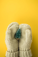 Female hands in white knitted winter mittens neatly hold a toy in the form of a green Christmas tree in the snow on a bright yellow background. View from above. Flat lay. Place for text. Copyspace.