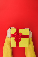 Female hands with manicure in an orange sweater hold gift shiny gold box on red background. Christmas and New Year holiday concept. Top view. Flat lay.