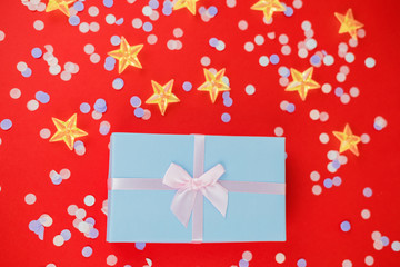 Festive Christmas and New Year background with blue gift box, yellow stars, multicolored confetti on bright red background. Top View. Flat lay. Concept of preparation for holidays.