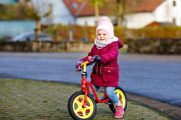 Cute little toddler girl riding on run balance bike to daycare, playschool or kindergarden. Happy healthy lovely baby child having fun with learning on leaner bicycle. Active kid on cold day outdoors.