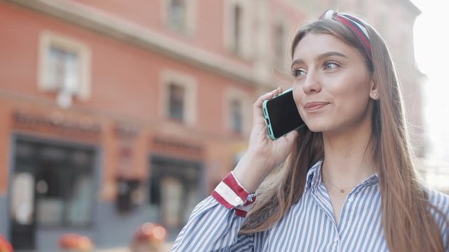 Young Attractive Girl Wearing in Striped Shirt Dialing Phone Number on Smartphone and Calling Talking and Smiling Standing on Street Outdoors Blurred Tramway on Background.