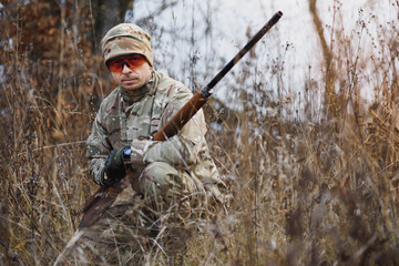 Fototapeta premium A male hunter with a gun while sitting takes aim at a forest. The concept of a successful hunt, an experienced hunter. Hunting the autumn season. The hunter has a rifle and a hunting uniform.