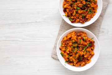 Homemade Chili Mac and Cheese with Parsley in white bowls over white wooden surface, top view. Flat lay, overhead, from above. Copy space.