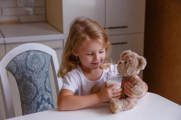 a little girl is sitting at the kitchen table at home, drinking milk and hugging teddy bear